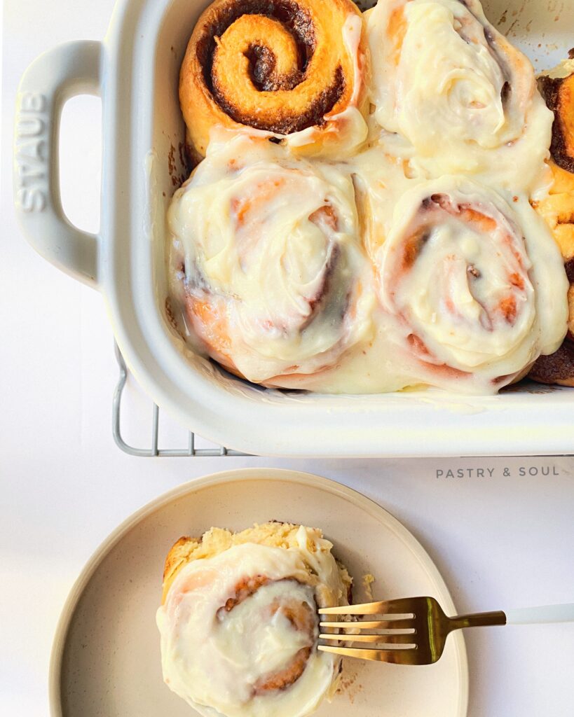 cinnamon rolls with cream cheese icing in a white baking dish and a white plate with a gold fork